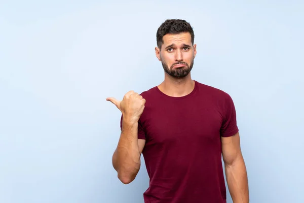 Hombre Guapo Sobre Fondo Azul Aislado Infeliz Apuntando Hacia Lado —  Fotos de Stock