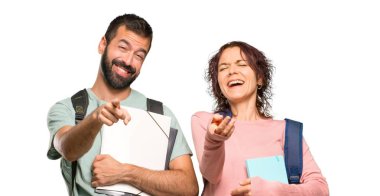 Sırt çantaları ve kitapları olan iki öğrenci birini parmakla gösteriyor ve izole beyaz arka plan da çok gülüyorlar.