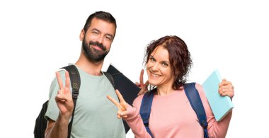 Sırt çantaları ve kitapları ile iki öğrenci gülümseyerek ve izole beyaz arka plan üzerinde iki eliyle zafer işareti gösteren