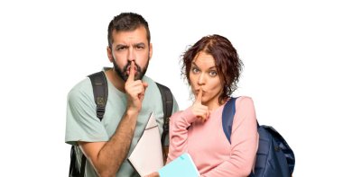 Sırt çantaları ve izole beyaz arka plan üzerinde ağız parmak koyarak sessizlik jest bir işareti gösteren kitap ile iki öğrenci