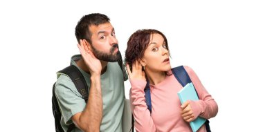 Sırt çantaları ve kitapları olan iki öğrenci izole beyaz arka plan üzerinde kulak elini koyarak bir şey dinlerken