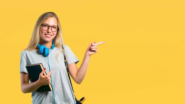 Student girl with glasses pointing finger to the side and presenting a product while smiling in a confident pose on yellow background