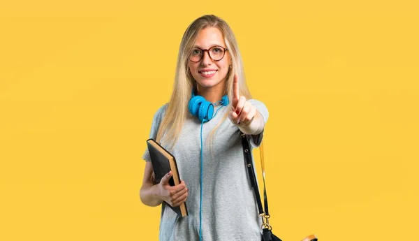 Student girl with glasses showing and lifting a finger in sign of the best on yellow background