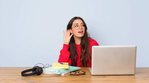 Teenager student girl studying in a table listening to something by putting hand on the ear
