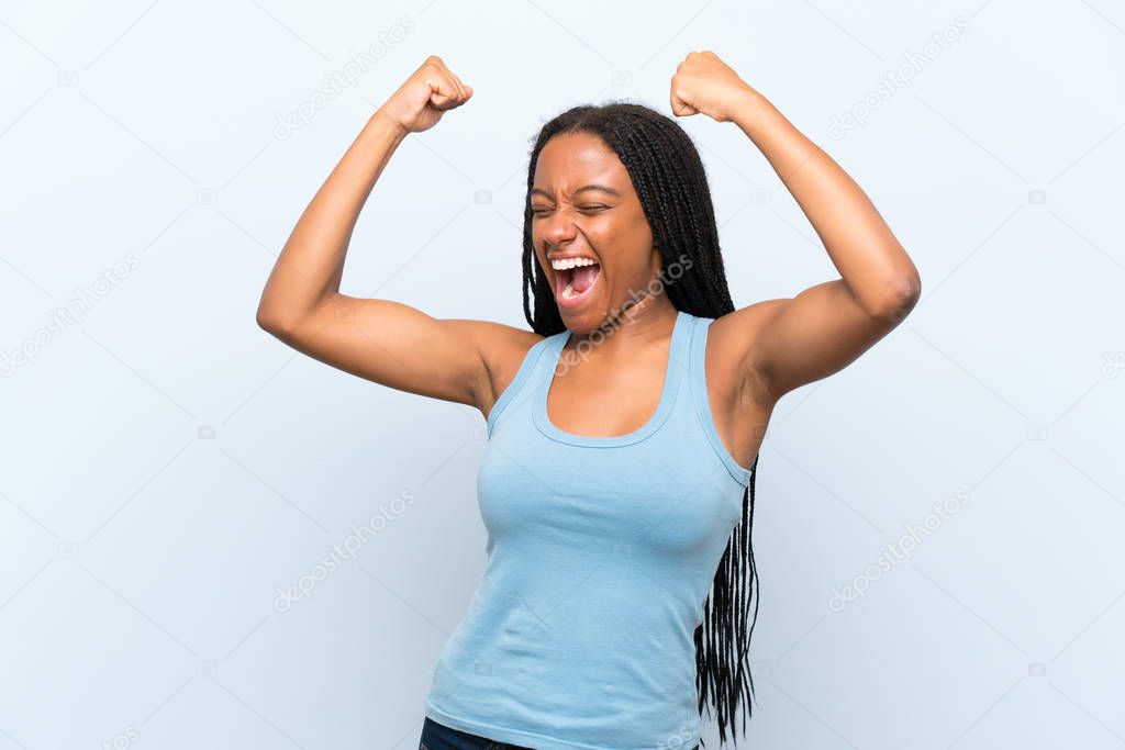 African American teenager girl with long braided hair over isolated blue background celebrating a victory