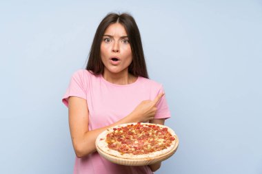 Izole mavi duvar üzerinde bir pizza tutan Pretty genç kız şaşırttı ve yan işaret