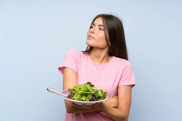 Young woman with salad over isolated blue background looking up while smiling