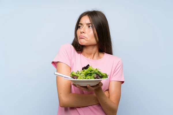 Young woman with salad over isolated blue background making doubts gesture while lifting the shoulders