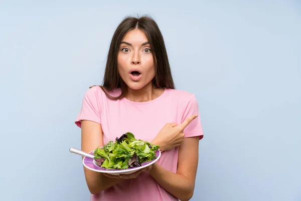 Young woman with salad over isolated blue background surprised and pointing side