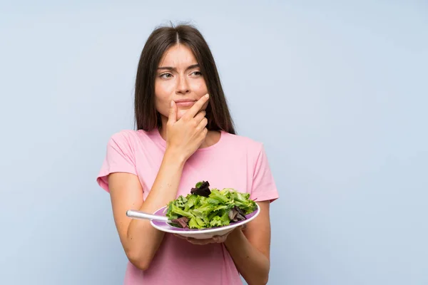 Young woman with salad over isolated blue background thinking an idea