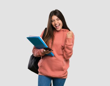 İzole gri arka plan üzerinde zafer konumunda telefon ile dizüstü tutan genç öğrenci kadın