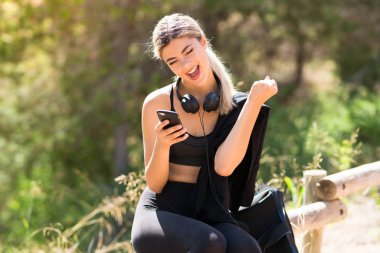 Genç spor kız zafer pozisyonunda telefon ile açık havada spor yapıyor