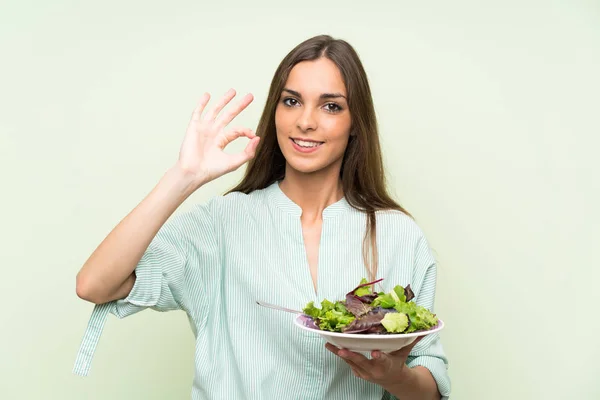 Young woman with salad over isolated green wall showing ok sign with fingers