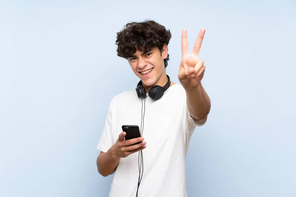 Young man listening music with a mobile over isolated blue wall smiling and showing victory sign