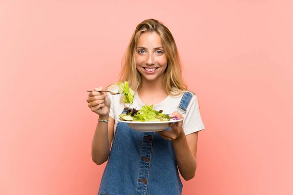 Gelukkige blonde jonge vrouw met salade over geïsoleerde roze muur — Stockfoto