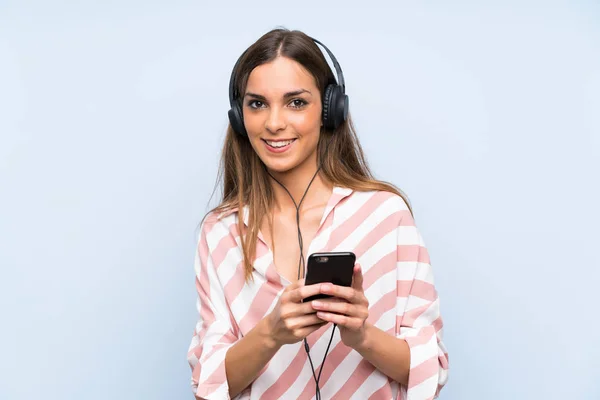 Mujer joven escuchando música con un móvil sobre una pared azul aislada —  Fotos de Stock