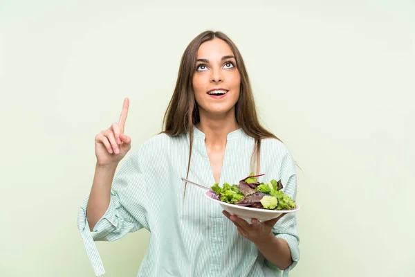 Young woman with salad over isolated green wall pointing up a great idea