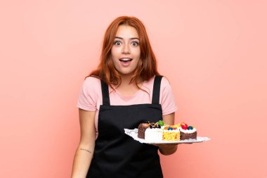Genç redhead kız sürpriz ve şok yüz ifadesi ile izole pembe arka plan üzerinde farklı mini kek çok tutan