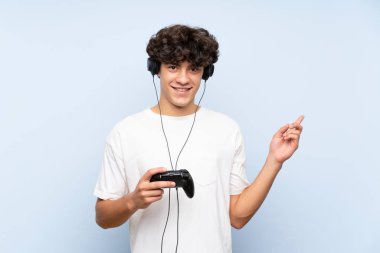 İzole mavi duvar üzerinde bir video oyunu denetleyicisi ile oynayan genç adam şaşırttı ve yan parmak işaret