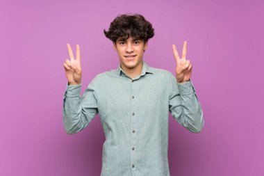 İzole edilmiş mor duvarlı genç adam iki eliyle zafer işareti gösteriyor.