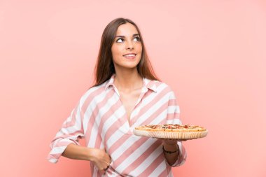 Genç kadın gülümserken yukarı bakıyor izole pembe duvar üzerinde bir pizza tutan