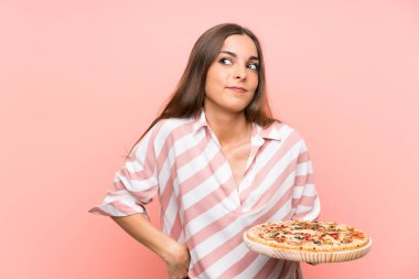 İzole pembe duvar üzerinde pizza tutan genç kadın omuzlarını kaldırırken şüphe jesti yapıyor