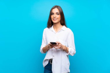 İzole edilmiş mavi arka planda genç bir kadın cep telefonuyla mesaj gönderiyor.