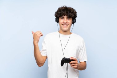 Bir video oyunu kontrolörüyle oynayan genç adam, izole edilmiş mavi duvarın üzerinde bir ürünü sunmak için yan tarafı işaret ediyor.