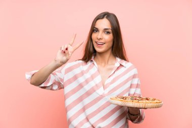 Genç kadın izole pembe duvar üzerinde bir pizza tutan gülümseyerek ve zafer işareti gösteren