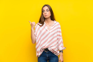 İzole edilmiş sarı arka planı olan genç bir kadın mutsuz ve bir tarafı işaret ediyor.