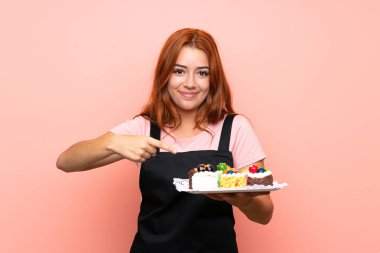 Genç redhead kız izole pembe arka plan üzerinde farklı mini kek çok tutan ve işaret