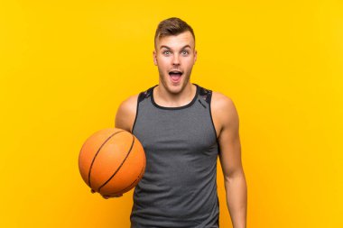 Genç yakışıklı sarışın adam sürpriz ve şok yüz ifadesi ile izole sarı arka plan üzerinde bir sepet topu tutan