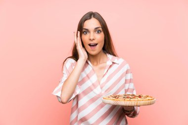 Genç kadın sürpriz ve şok yüz ifadesi ile izole pembe duvar üzerinde bir pizza tutan