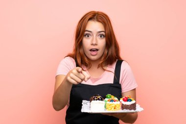Genç redhead kız izole pembe arka plan üzerinde farklı mini kek çok tutan şaşırttı ve ön işaret
