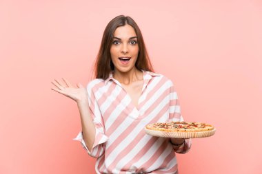 Genç kadın sürpriz yüz ifadesi ile izole pembe duvar üzerinde bir pizza tutan