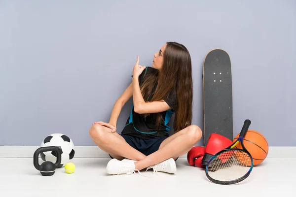 人差し指で後ろを指差して床に座っている若いスポーツ女性 — ストック写真