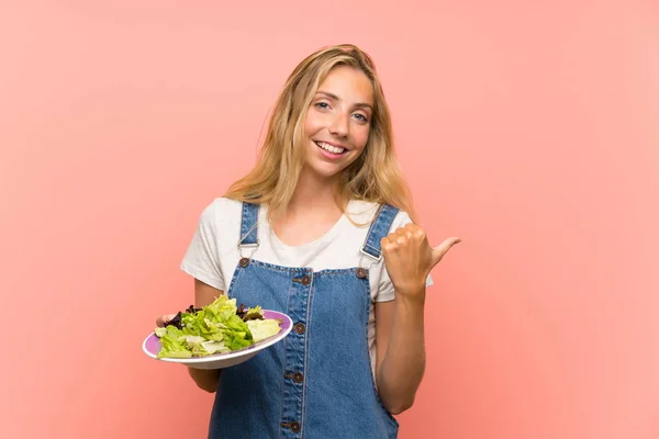 Blonde Jonge Vrouw Met Salade Geïsoleerde Roze Muur Wijzend Naar — Stockfoto