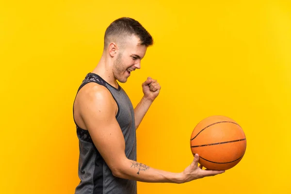 Young handsome blonde man holding a basket ball over isolated yellow background celebrating a victory
