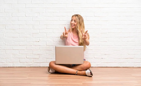 Young blonde student girl with a laptop on the floor smiling and showing victory sign