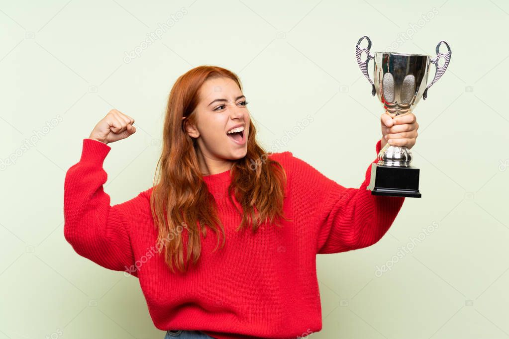 Teenager redhead girl with sweater over isolated green background holding a trophy