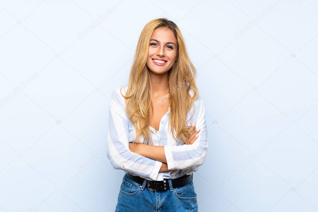 Young blonde woman over isolated blue background keeping the arms crossed in frontal position