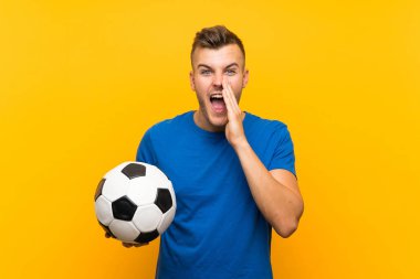 Genç yakışıklı sarışın adam ağzı geniş açık bağırarak izole sarı arka plan üzerinde bir futbol topu tutan