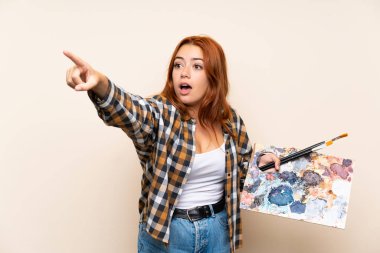 Genç kızıl saçlı kız uzak işaret izole arka plan üzerinde bir palet tutan