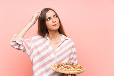 Genç kadın izole pembe duvar üzerinde şüpheler ve karıştırılmış yüz ifadesi ile bir pizza tutan