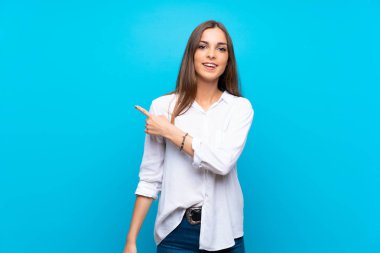 İzole edilmiş mavi arka planda genç bir kadın bir ürünü sunmak için kenarı işaret ediyor.