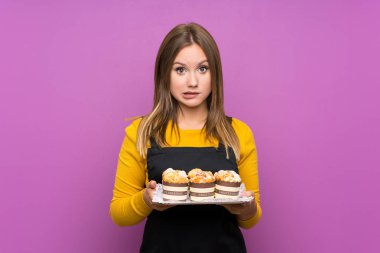 Genç kız izole mor arka plan üzerinde farklı mini kek çok tutan şüpheler ve yüz ifadesi karıştırarak