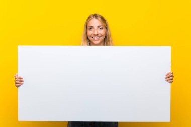 Bir kavram eklemek için boş bir beyaz pankart tutan izole sarı arka plan üzerinde Blonde genç kadın