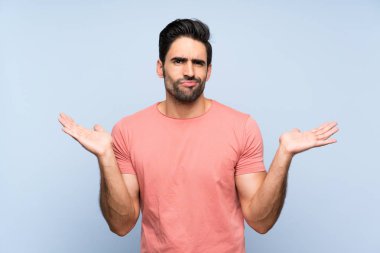Handsome young man in pink shirt over isolated blue background having doubts while raising hands