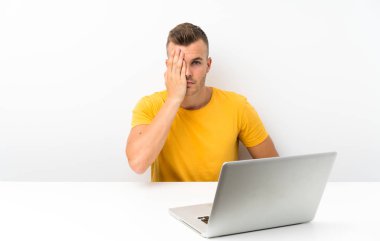 Young blonde man in a table with a laptop covering a eye by hand