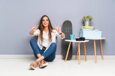 Yerde oturmuş gülümseyen ve zafer işareti gösteren genç bir kadın.